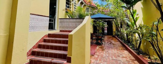 Yellow building exterior with red-tiled steps and patterned wall tiles, small outdoor seating area under a blue umbrella amid tropical plants.