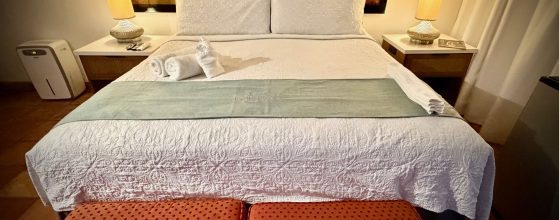 Hotel-style bedroom with a white quilted bed, two white pillows, rolled towels at the foot, and warm lamps on nightstands beside a large artwork behind the bed.